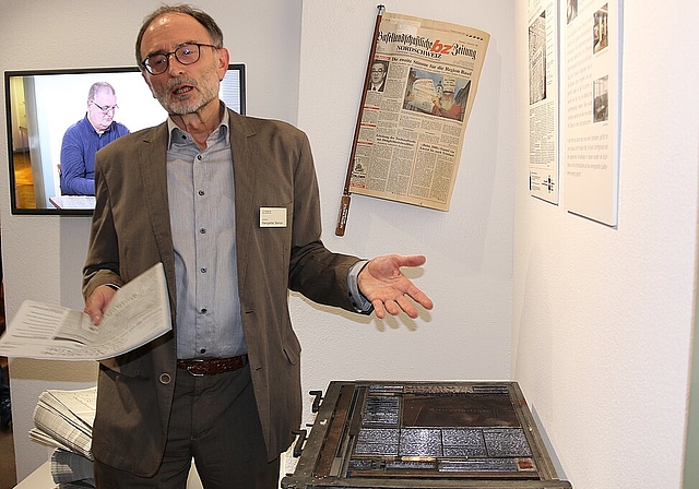 Zeitdokumente: Hanspeter Steiner, Leiter Ausstellung, präsentiert die Sonderzeitung, deren Frontseite nach altem Druckverfahren mit Bleisatz und Druckform hergestellt wurde. Auf dem Bildschirm im Hintergrund berichtet Thomas Immos über seine Zeit a